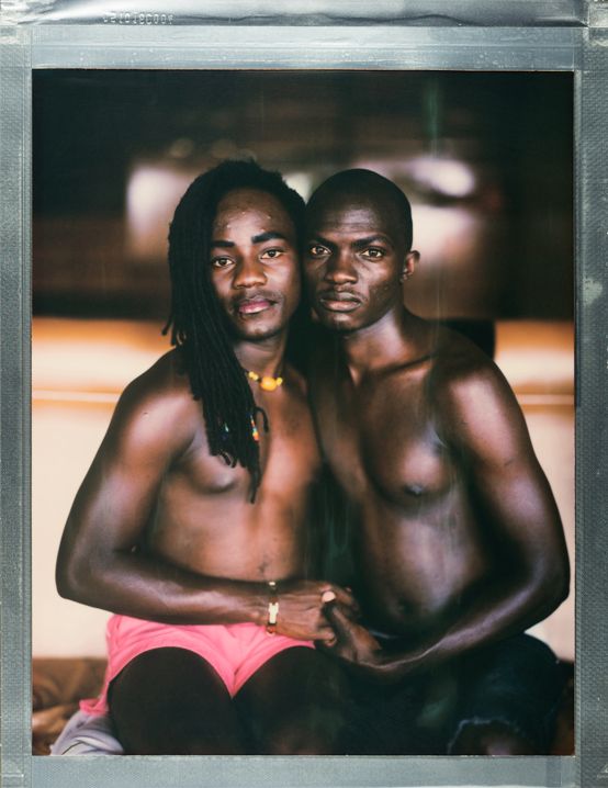 A posed portrait of Lucky (right) and John (left), Ugandan refugees living in Nairobi. Lucky and John lived together in Uganda - until John’s parents found out they were in a relationship and attacked Lucky.They hid with a friend and saved enough money to flee to Kenya. They were registered separately as refugees and they were able to find some sanctuary in Nature Network. “The life now in Nairobi, because of the Nature Network we have, the little money we are getting, it help me someway, somehow, and the Nature Network come in, they do pay us rent here, they buy us food.” Faith has been an important part of keeping them strong through their trials.
“If it wasn't God's help, we would have already died, because I remember the time when the parents came to attack him [Lucky], and then, they wanted to kill him, if it was not God, he would have already died, but God knows us, God loves us, so he managed to protect us all the way from Uganda up to here, we are together.” Kenya, October 2017. 
Nature Network is a Nairobi based organization providing LGBTQI+ refugees in Kenya with support through safe temporary housing, health services, food and security. Nature Network has advocated to police over 50 times, responding to hate crimes, and runs a WhatsApp group of safety tips. Refugees supported have come from Uganda, Somalia, Burundi, Rwanda and Sudan. 
Stigma, discrimination and violence based on sexual orientation means that access to HIV services is yet another challenge for this community. As a result, LGBTQI+ people in Africa are 19 times more likely to be living with HIV, with prevalence rates in many countries exceeding 10-20%. To respond to this, the Elton John AIDS Foundation (EJAF) built a quick, nimble, and easily accessible $10m fund which can get money to the most effective grassroots organisations doing some of the most important work among the most-at-risk LGBT groups in Sub-Saharan Africa.  A Rapid Response mechanism administered by the International HIV / AIDS Alliance quickly disburses smaller sums to respond to emergencies where LGBT people are in jeopardy. The fund is active in 30 countries and Nature Network in Kenya is one project that has received the fund.  
Robin Hammond/NOOR for Witness Change